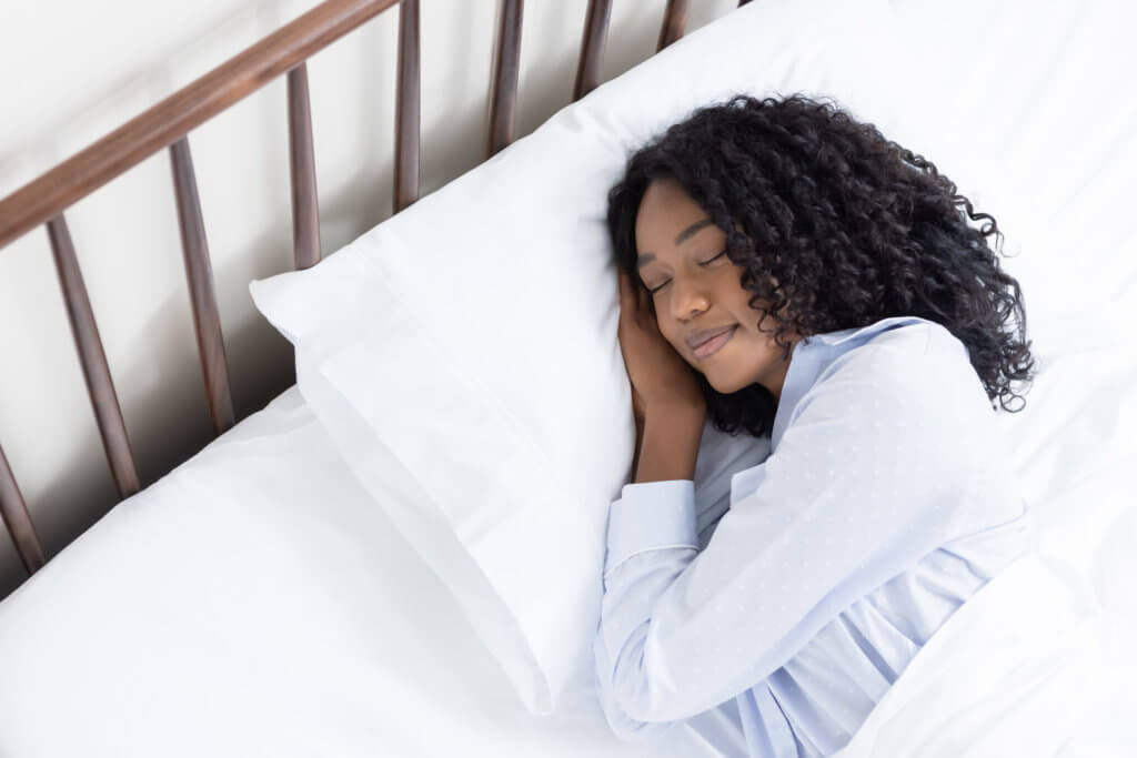 A woman places her hands under head on a plush, white pillow, covered in her sateen cotton sheets.